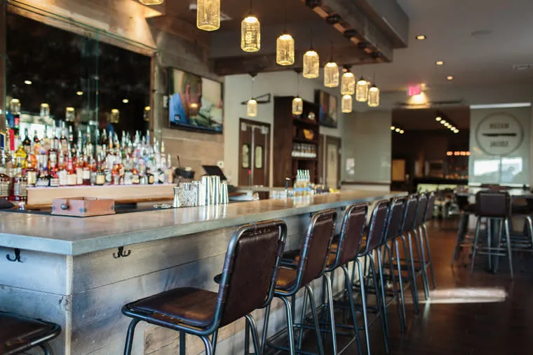 A modern bar with a long wooden counter, high bar stools, hanging jar lights, and shelves of liquor bottles. The space appears cozy and inviting, with warm lighting and a few tables in the background.