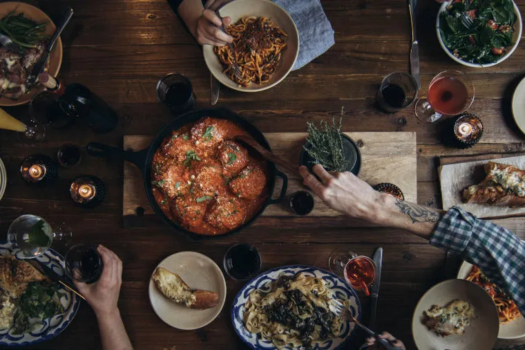 A cozy dinner table scene with several hands reaching for Italian food, including pasta, salad, bread, wine, and a large skillet of meatballs in tomato sauce, all surrounded by lit candles.