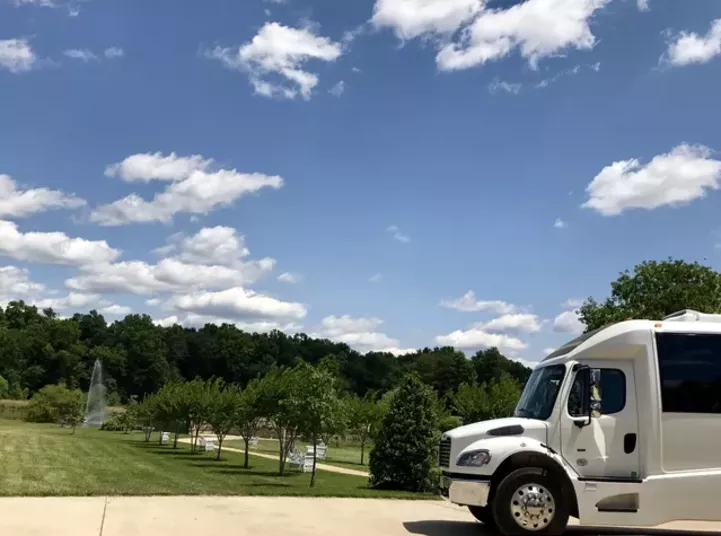 A large white truck is parked on a driveway under a blue sky with scattered clouds. Green trees and a grassy area with a small fountain are visible in the background.