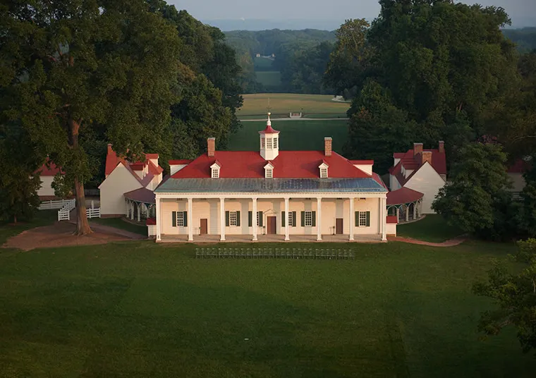 A large, white historic mansion with a red roof and columned porch sits surrounded by green lawns and trees, with smaller outbuildings nearby and a scenic landscape in the background.