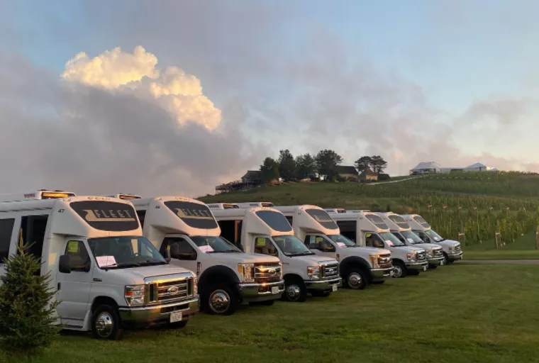 A row of white shuttle buses is parked on grass near a hillside with trees and buildings in the background under a partly cloudy sky at sunset.