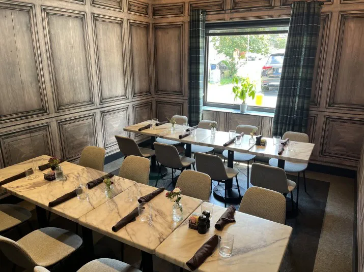 A small, elegant dining room with two marble tables set for a meal. Beige chairs surround the tables, which are adorned with glassware, napkins, and small flower vases. A window lets in natural light.