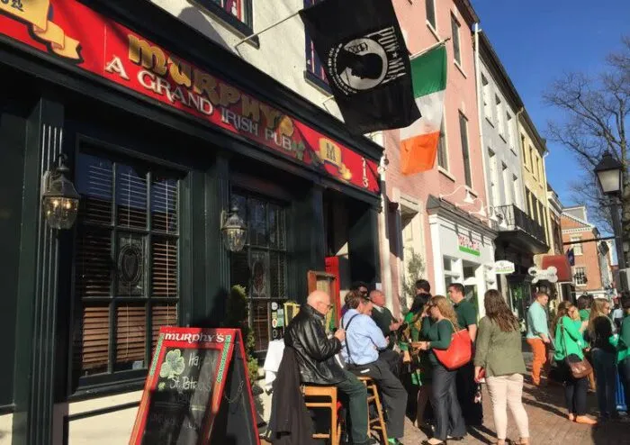 People gather outside Murphy’s Irish Pub, beneath Irish and POW/MIA flags, on a sunny day. A signboard is visible near the entrance. The pub has a red facade and the street is lined with other buildings.