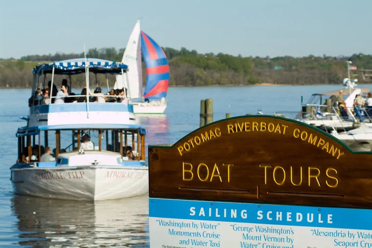 A Potomac Riverboat Company sign promotes boat tours as a white tour boat and a colorful sailboat travel on the calm river in the background under a clear sky.