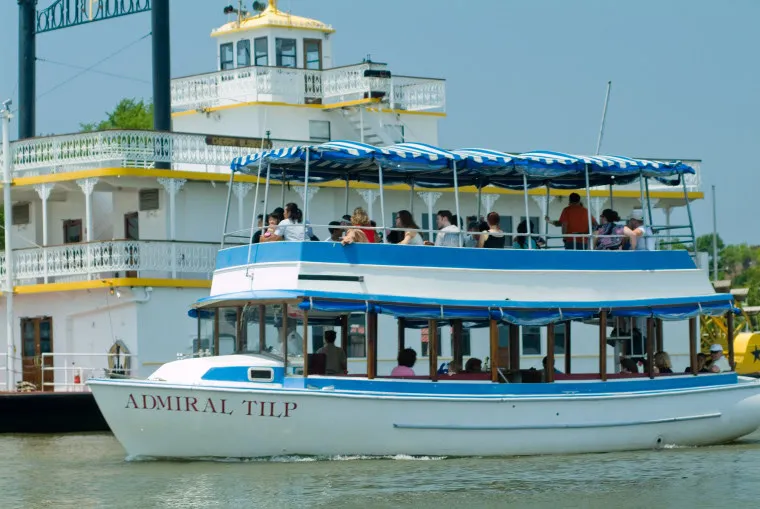 A white and blue tour boat named “Admiral Tilp” with passengers on board cruises on a river. In the background, a larger riverboat with yellow trim is docked. The weather is sunny and calm.