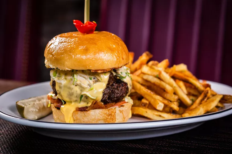 A cheeseburger with a glossy bun, melted cheese, and lettuce, served with a pickle spear and a pile of golden French fries on a white plate. A small red garnish sits on top of the burger bun.