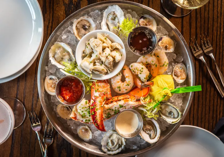 A seafood platter on ice with oysters, shrimp, crab meat, a lobster claw, cocktail sauce, creamy sauce, lemon, and greens, served on a wooden table with plates, forks, and drinks nearby.