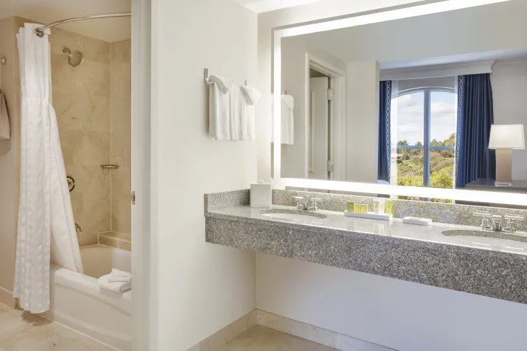A hotel bathroom with a double-sink vanity, large illuminated mirror, and a bathtub with a shower curtain. White towels hang on the wall, and a window reveals a scenic outdoor view.