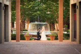 A man in a suit kneels and proposes to a woman in a white dress near a fountain in a courtyard, surrounded by trees and brick columns.
