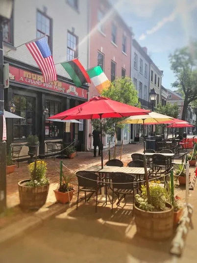 Outdoor seating area with red and yellow umbrellas on a brick-paved street, outside a restaurant displaying American, Irish, and Italian flags. Sunlight creates a warm, inviting atmosphere.