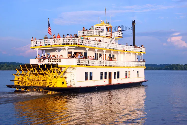 A white and yellow paddlewheel riverboat named Cherry Blossom carries passengers on a calm river, with an American flag flying at the stern and trees visible in the background under a partly cloudy sky.