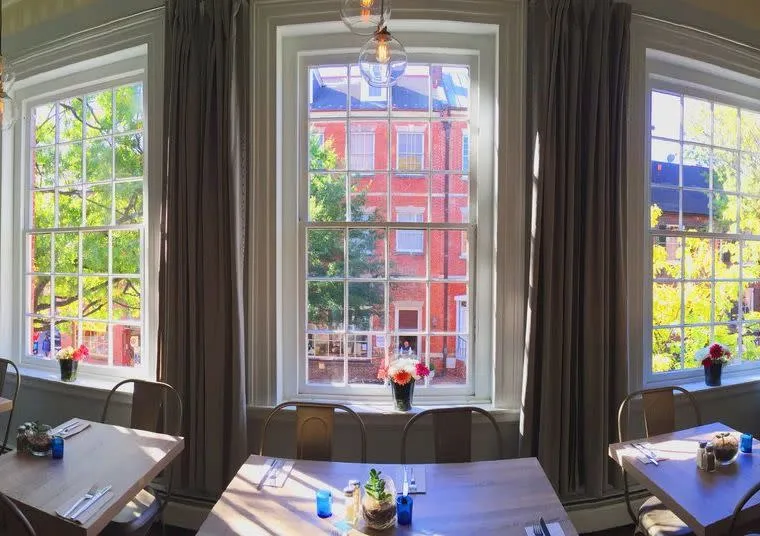 Daylight streams through three large windows with white frames and beige curtains, overlooking a red-brick building and trees. Tables with chairs, place settings, and small flower vases are visible in the cozy, well-lit room.