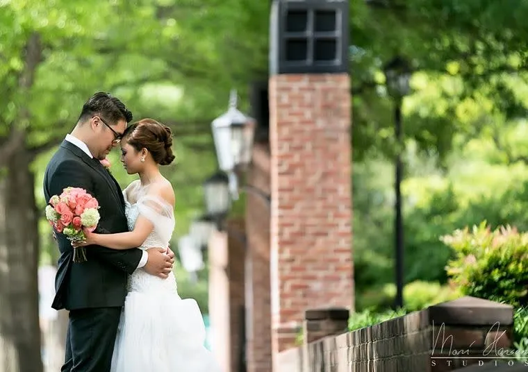 A bride and groom embrace outdoors, standing close with foreheads touching. The bride holds a bouquet of pink flowers and wears a white dress, while the groom is in a black suit. Green trees and brick walls are in the background.
