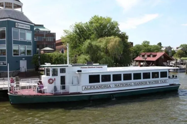 A white and green water taxi labeled Alexandria - National Harbor Water Taxi is docked by a riverside with trees and buildings in the background. Several people are seated on the boat.