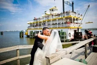 A bride and groom share a kiss on a dock, with a large, yellow and white riverboat full of passengers in the background on a sunny day.