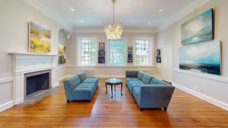 A bright living room with wooden floors, two blue sofas facing each other around a round glass coffee table, a fireplace on the left, large windows, and several landscape paintings on the walls.