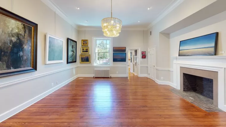 A spacious art gallery room with wooden floors, cream-colored walls, a chandelier, and various paintings hung on the walls. A large window and a fireplace are visible, letting in natural light.