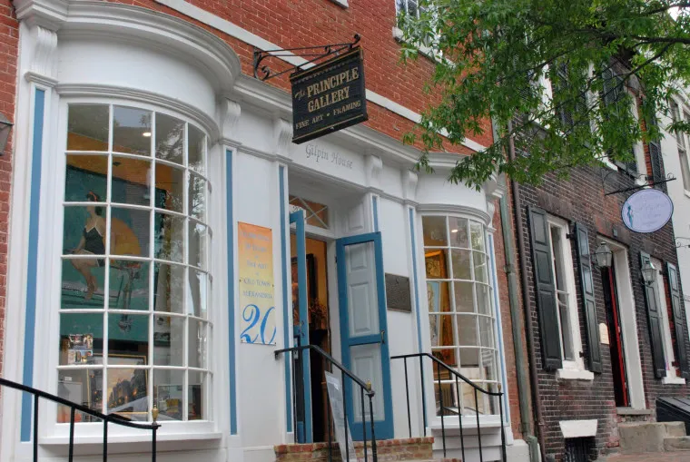 A brick building with large bay windows and an open blue door houses the Principle Gallery. The gallery sign hangs above the entrance, and artwork is visible through the windows. Trees line the sidewalk in front.