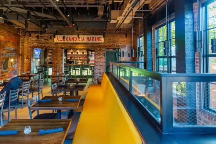 A bright, modern restaurant with exposed brick walls, large windows, wooden tables, a long yellow cushioned bench, and a bar area. A sign reads “ALEXANDRIA MARINE.” One person sits at a table.