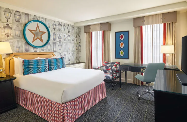 A brightly lit hotel room features a queen bed with a patterned headboard and blue pillows, a desk with a blue chair, patterned armchair, and wall art, including a large star and colorful framed circles.
