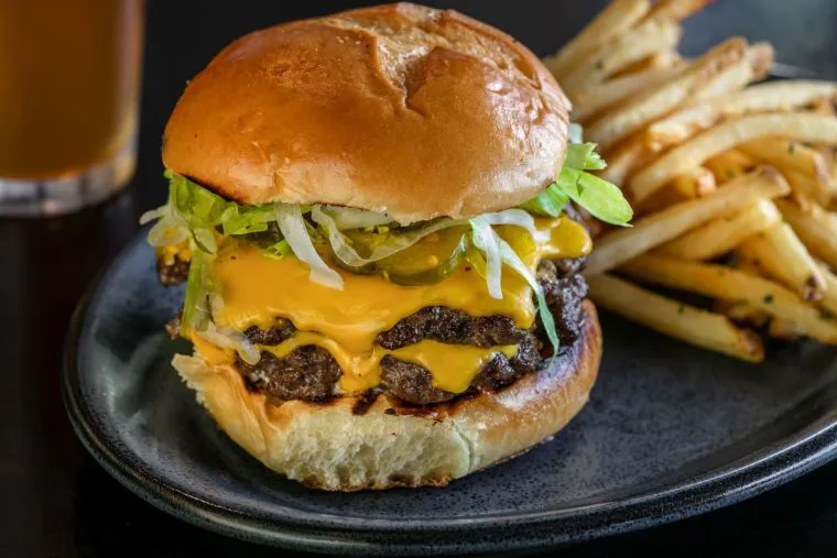 A cheeseburger with lettuce, pickles, and two beef patties is served on a bun next to a pile of seasoned French fries on a dark plate.