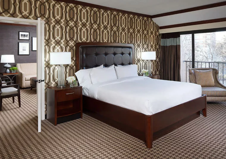 Modern hotel room with a large bed, dark wood furniture, geometric-patterned wallpaper, two bedside lamps, a sitting area with an armchair, and a sliding glass door leading to a balcony.