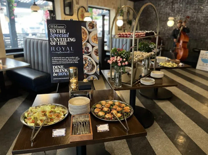 A buffet setup in a restaurant features salads, pastries, and desserts on tables, with flowers and a welcome sign. In the background, a musician plays a double bass near a window.