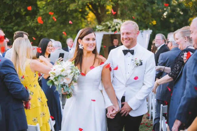 A bride and groom walk down the aisle outdoors, smiling and holding hands, surrounded by guests tossing red flower petals in celebration. The bride holds a bouquet and wears a white dress; the groom wears a white tuxedo jacket.