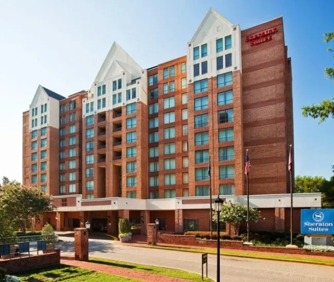 A multi-story brick hotel building with white trim, balconies, and peaked roofs, with the Sheraton Suites sign and two flagpoles in front, surrounded by greenery under a clear blue sky.