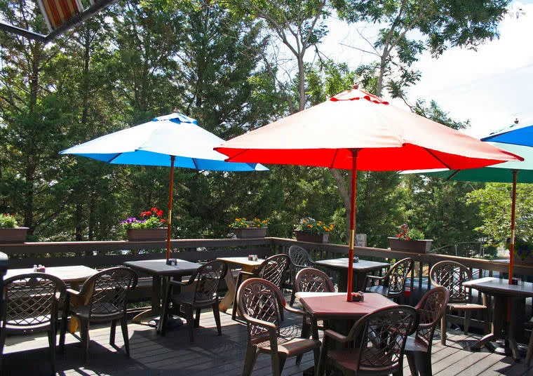 Outdoor patio with tables and chairs, shaded by colorful blue and red umbrellas. Surrounded by green trees and flower boxes along the wooden railing, creating a relaxed, sunny atmosphere. No people are present.