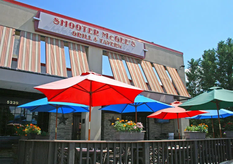 Outdoor patio at Shooter McGee’s Grill & Tavern, featuring colorful umbrellas, flower boxes, and striped awnings on the building, with trees and a clear blue sky in the background.