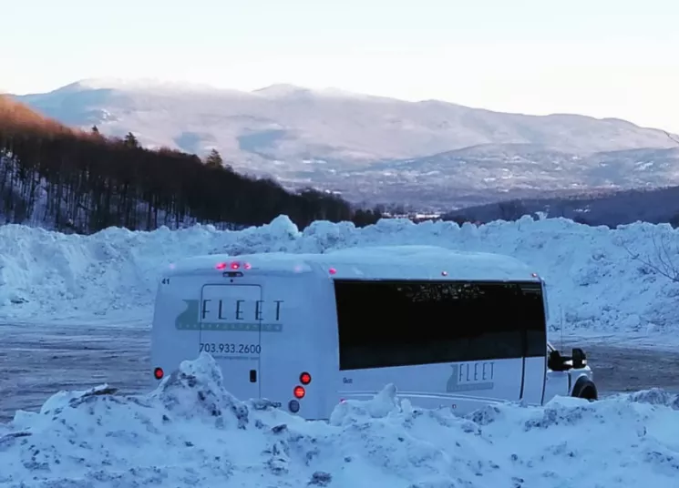 A white shuttle bus with “FLEET” branding is parked in a snowy lot surrounded by large snowbanks. Mountains covered in snow and trees are visible in the background under a clear sky.