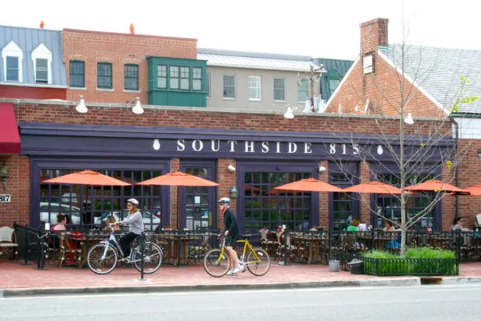 People ride bicycles past an outdoor patio with orange umbrellas at a restaurant called Southside 815 on a city street lined with brick buildings.