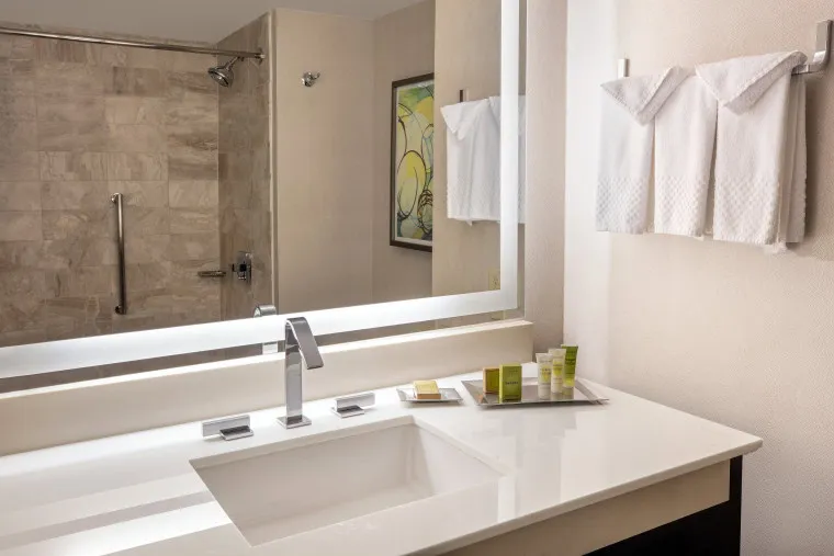 Modern hotel bathroom with a white countertop, rectangular sink, and a large backlit mirror. Towels are neatly hung and folded on the wall, and toiletries are arranged on a tray. Shower stall with glass door is in the background.