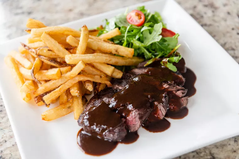 A square white plate with steak topped in dark sauce, golden French fries, and a side salad with greens and cherry tomatoes, set on a granite countertop.