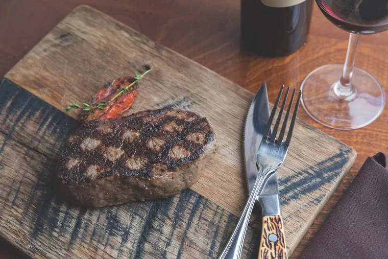 A grilled steak with sear marks is served on a wooden cutting board with a roasted tomato garnish. A fork and knife rest beside the steak, with a glass of red wine and a wine bottle nearby.