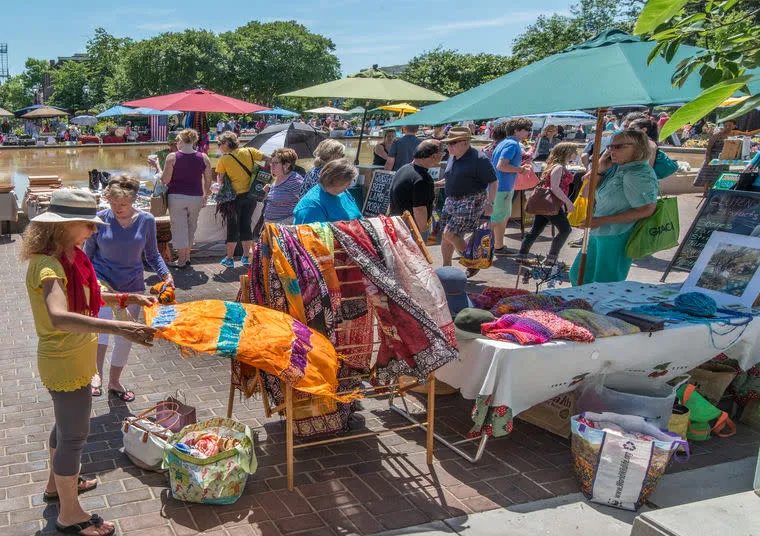 People browse colorful handmade textiles at an outdoor market on a sunny day. Various stalls, umbrellas, and trees are visible, with shoppers examining goods and socializing.