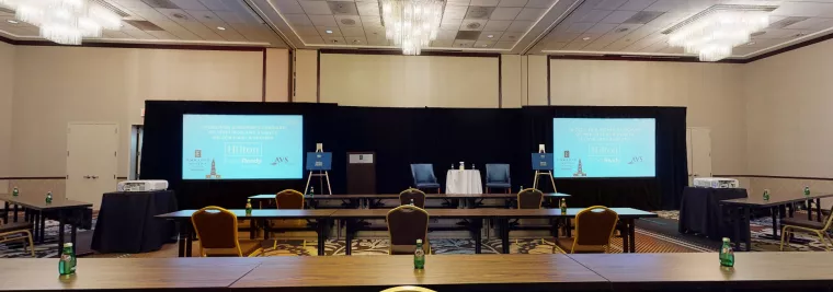 A conference room with chandeliers, rows of tables and chairs, two large screens displaying event information, and bottled water placed on tables. The setup faces a stage with two chairs and a small table.
