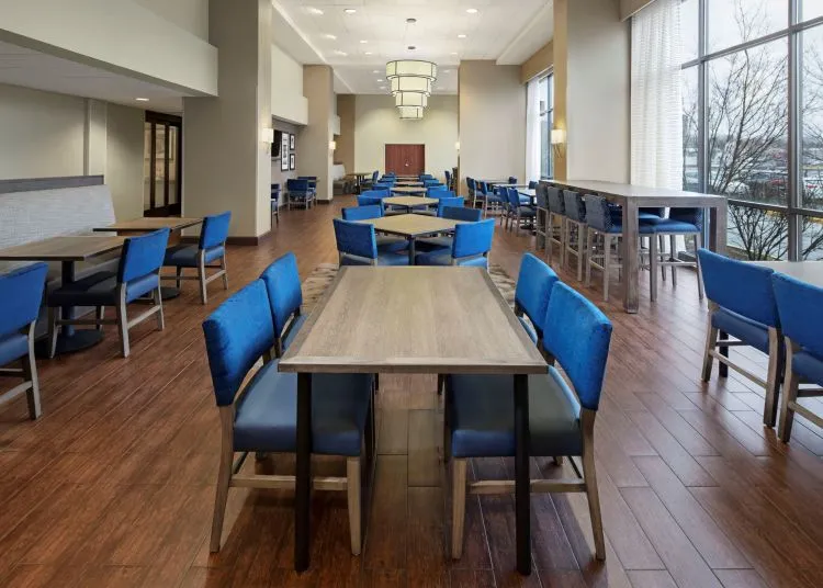 Modern dining area with wooden floors, light-colored walls, and large windows. Blue cushioned chairs are arranged around wooden tables, and natural light fills the spacious, clean room.