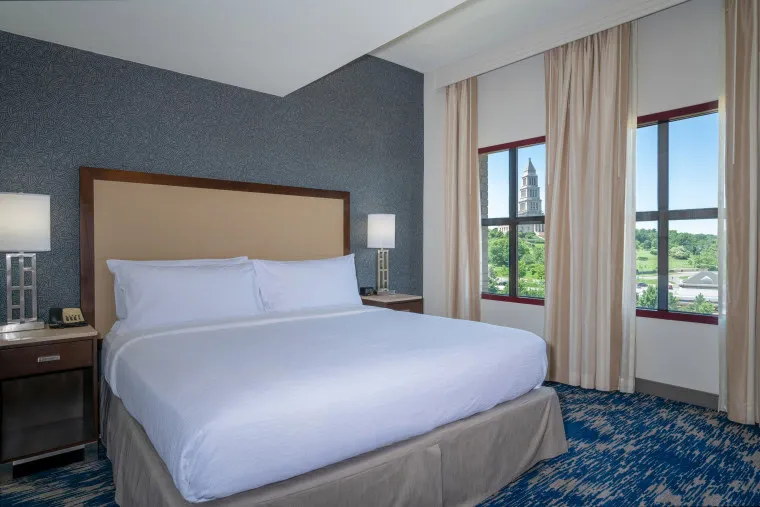 A hotel room with a large neatly made bed, two bedside tables with lamps, and large windows with beige curtains. Outside, a tower and greenery are visible in the distance.