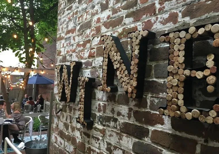 Large letters spelling WINE are mounted on a brick wall and filled with wine corks. In the background, people sit at tables in an outdoor patio strung with lights, beneath trees and umbrellas.