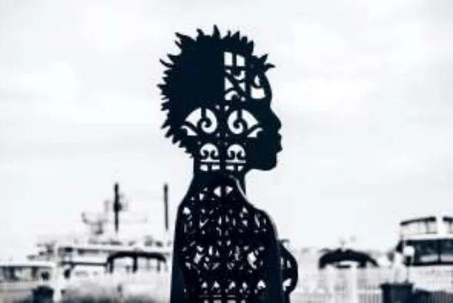 Silhouette of a decorative metal sculpture shaped like a persons side profile, with intricate patterns, set against a blurred cityscape background.