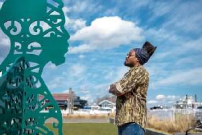 A man wearing glasses and a patterned shirt stands outdoors, facing a green decorative metal sculpture shaped like a persons profile, with blue sky and buildings in the background.