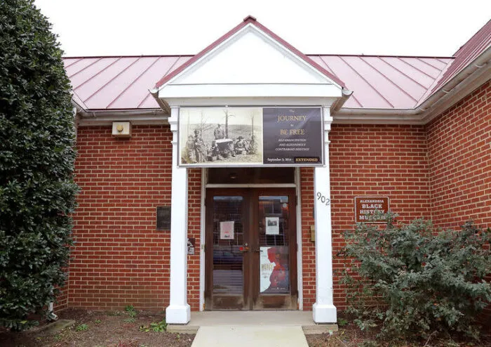 A red brick building with white columns at the entrance, displaying a banner above the door. A sign on the right reads “Alexandria Black History Museum.” Shrubs line the walkway to the entrance.