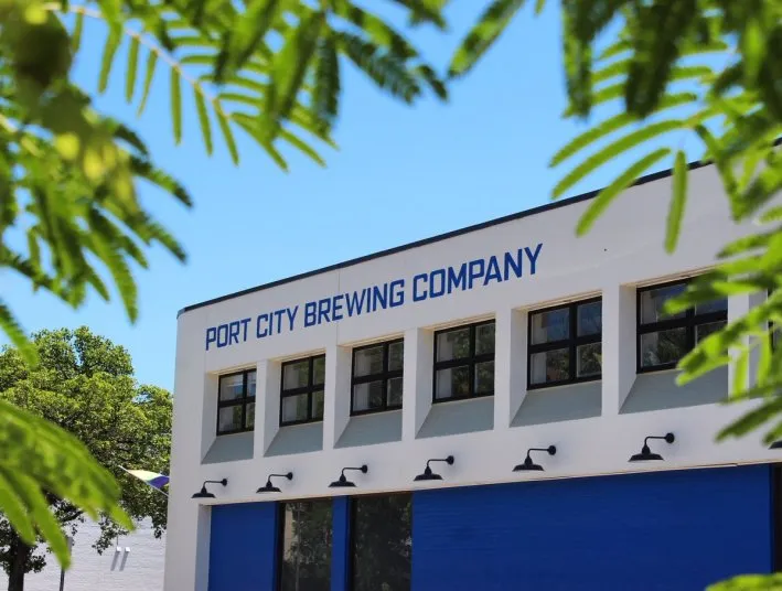 A white and blue building with Port City Brewing Company written on the side, partially framed by green leaves, under a clear blue sky.