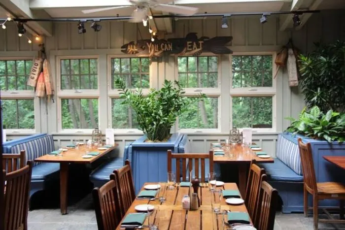 A cozy restaurant dining area with wooden tables, blue cushioned booths, set tables, plants, windows showing greenery outside, and a sign above reading All You Can Eat.