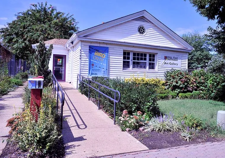 A small white art gallery building with a ramp, surrounded by lush gardens and colorful flowers. A blue banner hangs by the entrance, and a red mailbox stands by the walkway leading to the door.