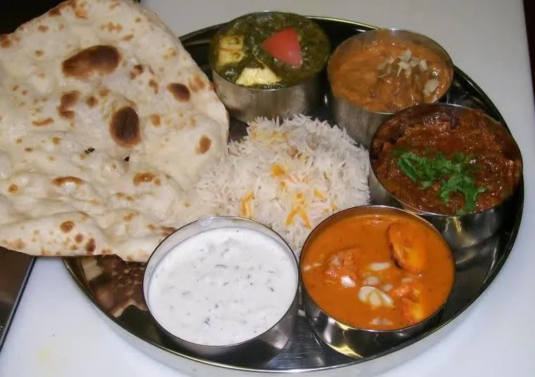 A metal plate with naan bread, basmati rice, and five bowls containing Indian dishes: palak paneer, butter chicken, a curry with cilantro, raita, and another curry, all served on a white surface.