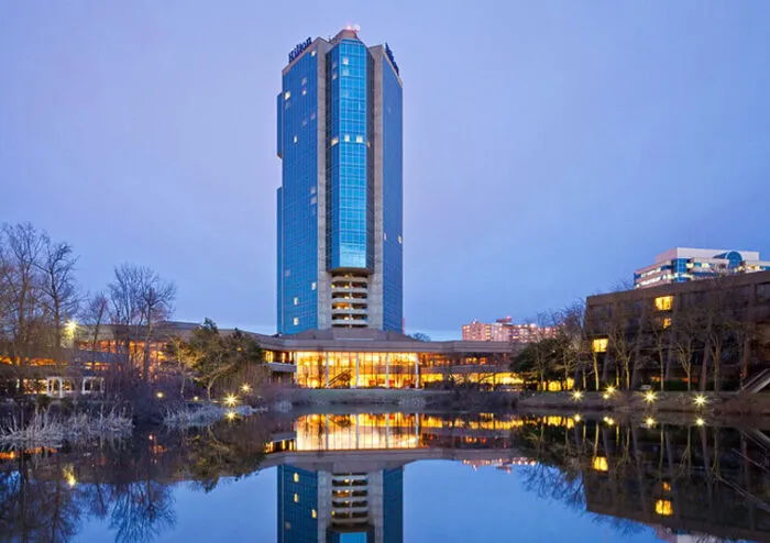 A tall modern hotel tower with blue-tinted windows rises above a landscaped area and reflects in a calm pond at dusk, with warm lights glowing from the building.