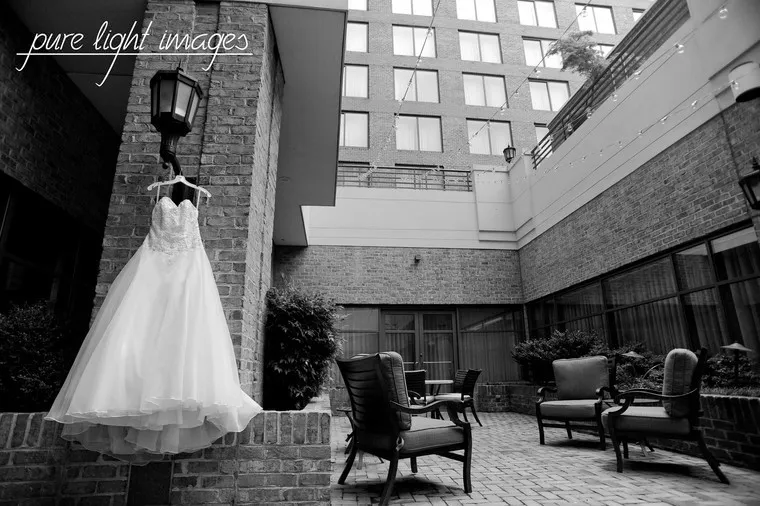 A wedding dress hangs on a brick wall in a courtyard with outdoor seating, surrounded by tall buildings. The image is black and white, with “pure light images” written in the top left corner.